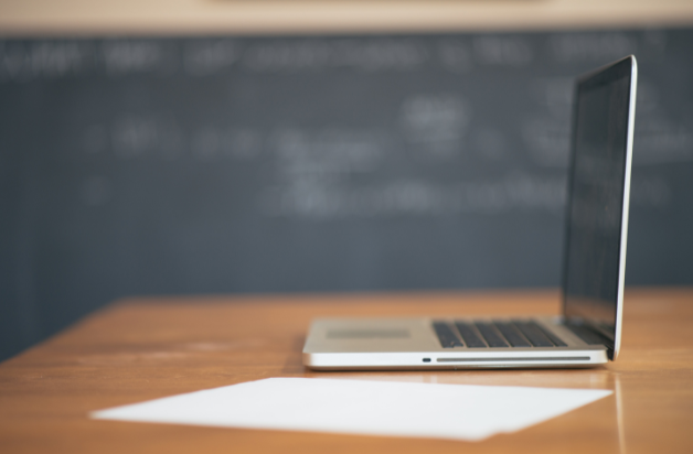 A picture of a laptop and a piece of paper with a blackboard in the background. 