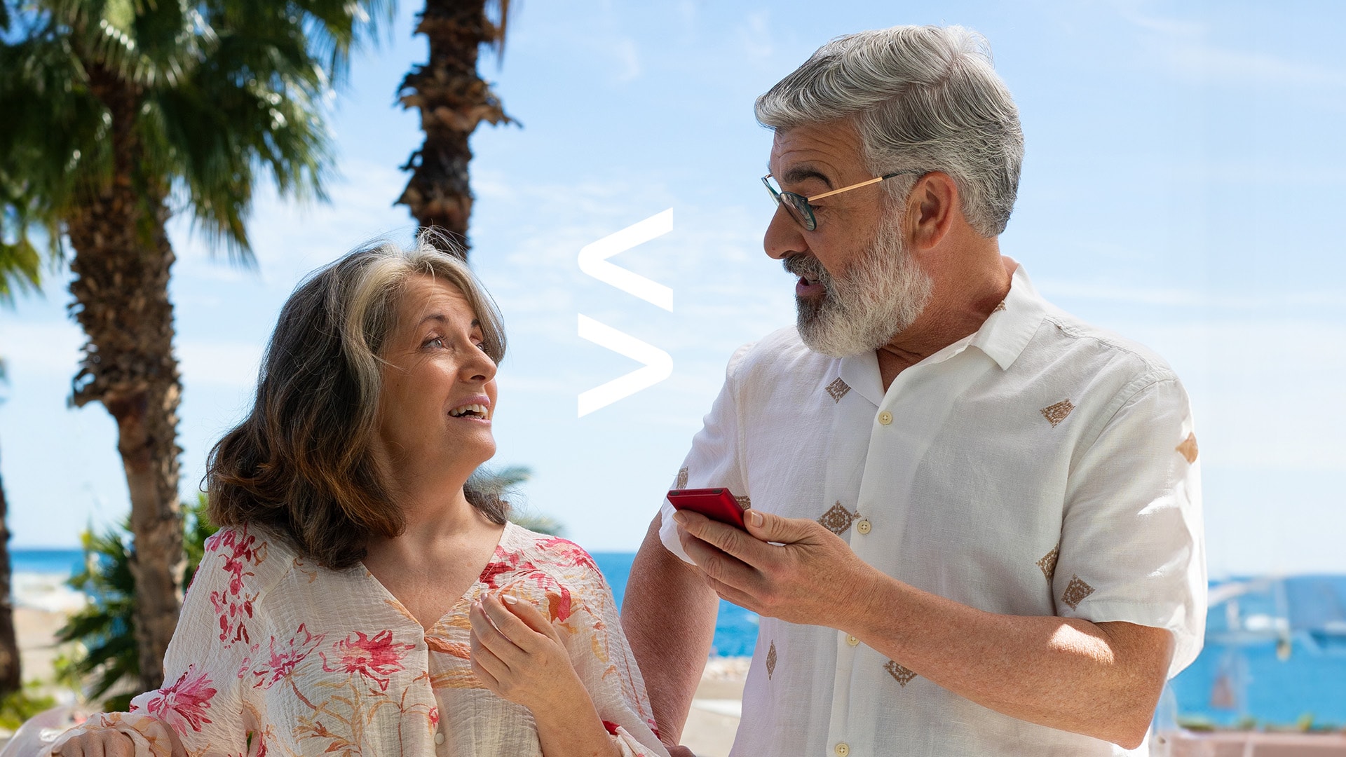 An elderly couple stands outdoors by the sea, using a Vasco Translator V4 device to communicate with each other, enjoying a conversation under the palm trees.