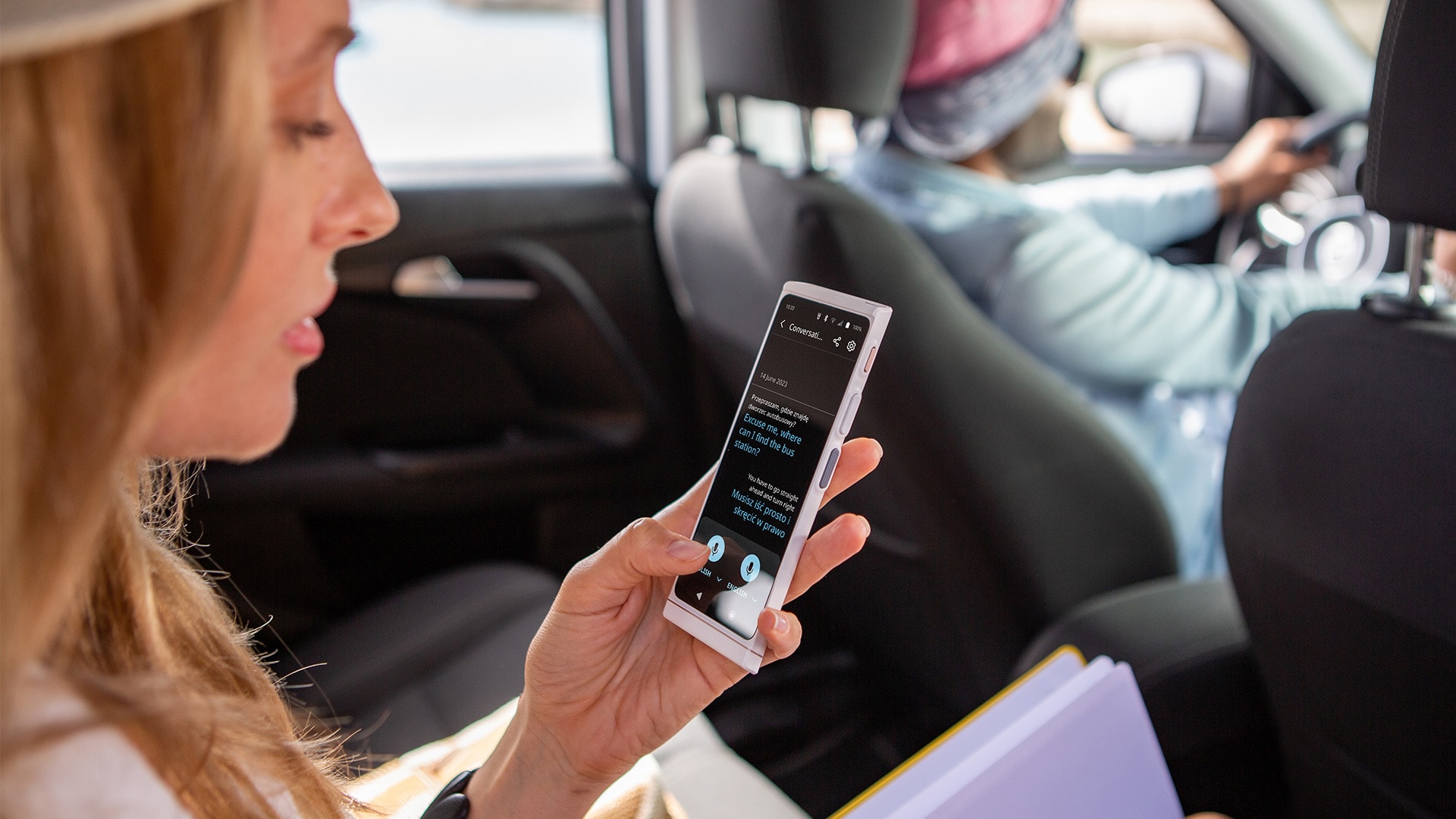 A woman sits in the backseat of a car, holding a Vasco Translator V4 device, likely using it to communicate with the driver.