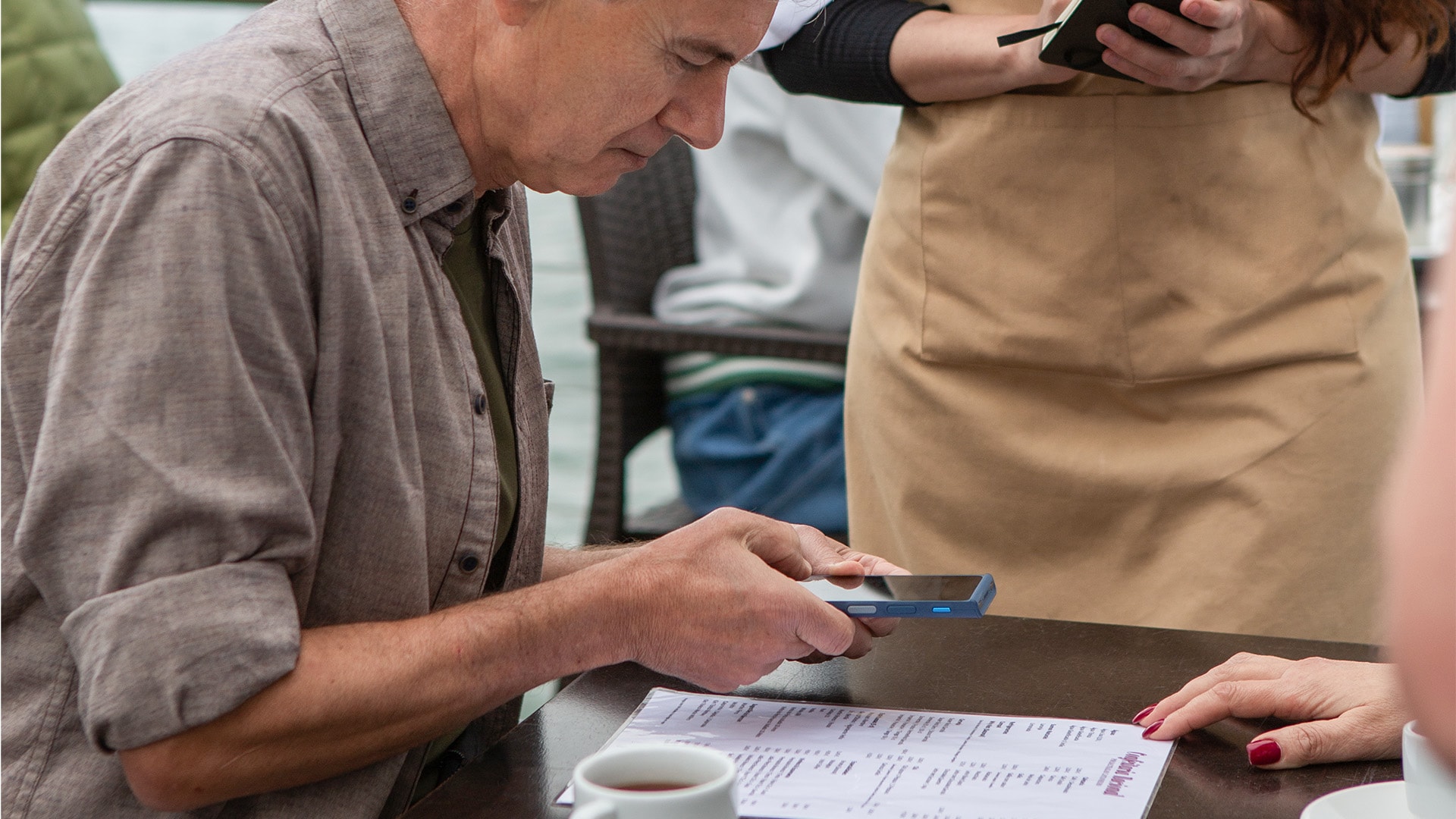 A man seated at a table is using a Vasco Translator V4 to translate a menu, while a woman stands nearby with another device.