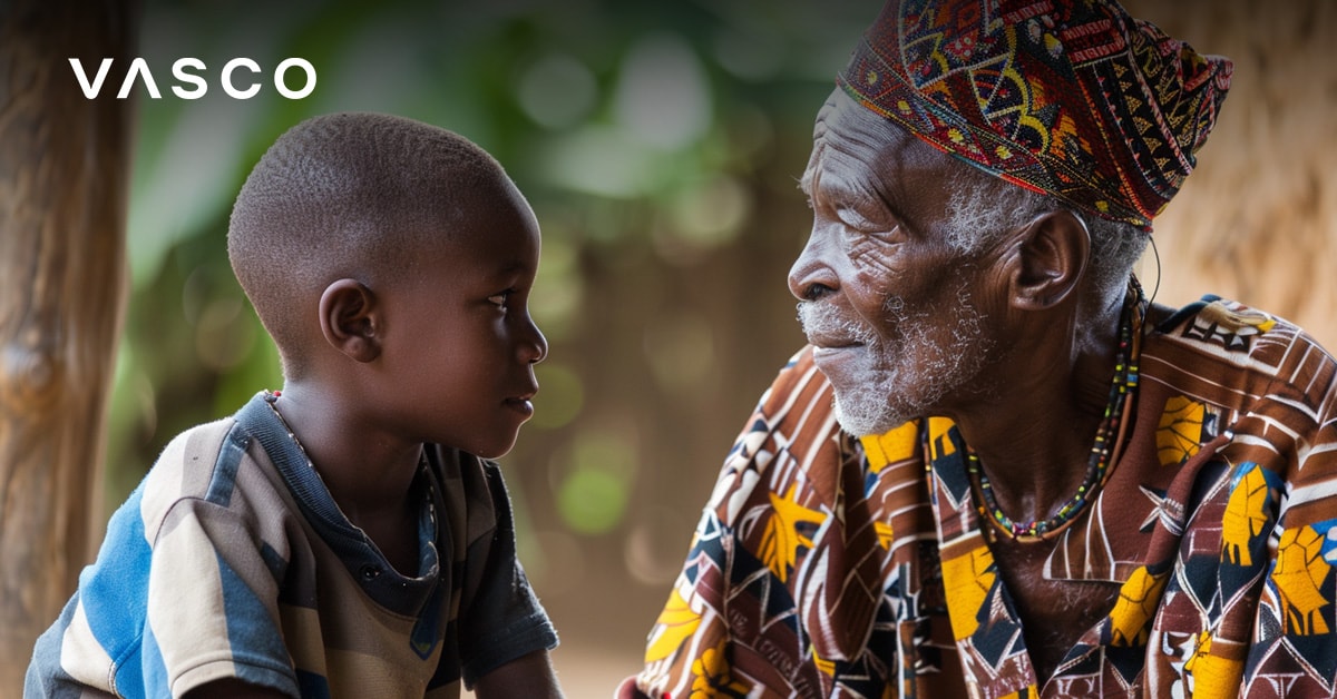 An elderly man in colorful traditional attire sitting and talking with a young boy, both gazing intently at each other