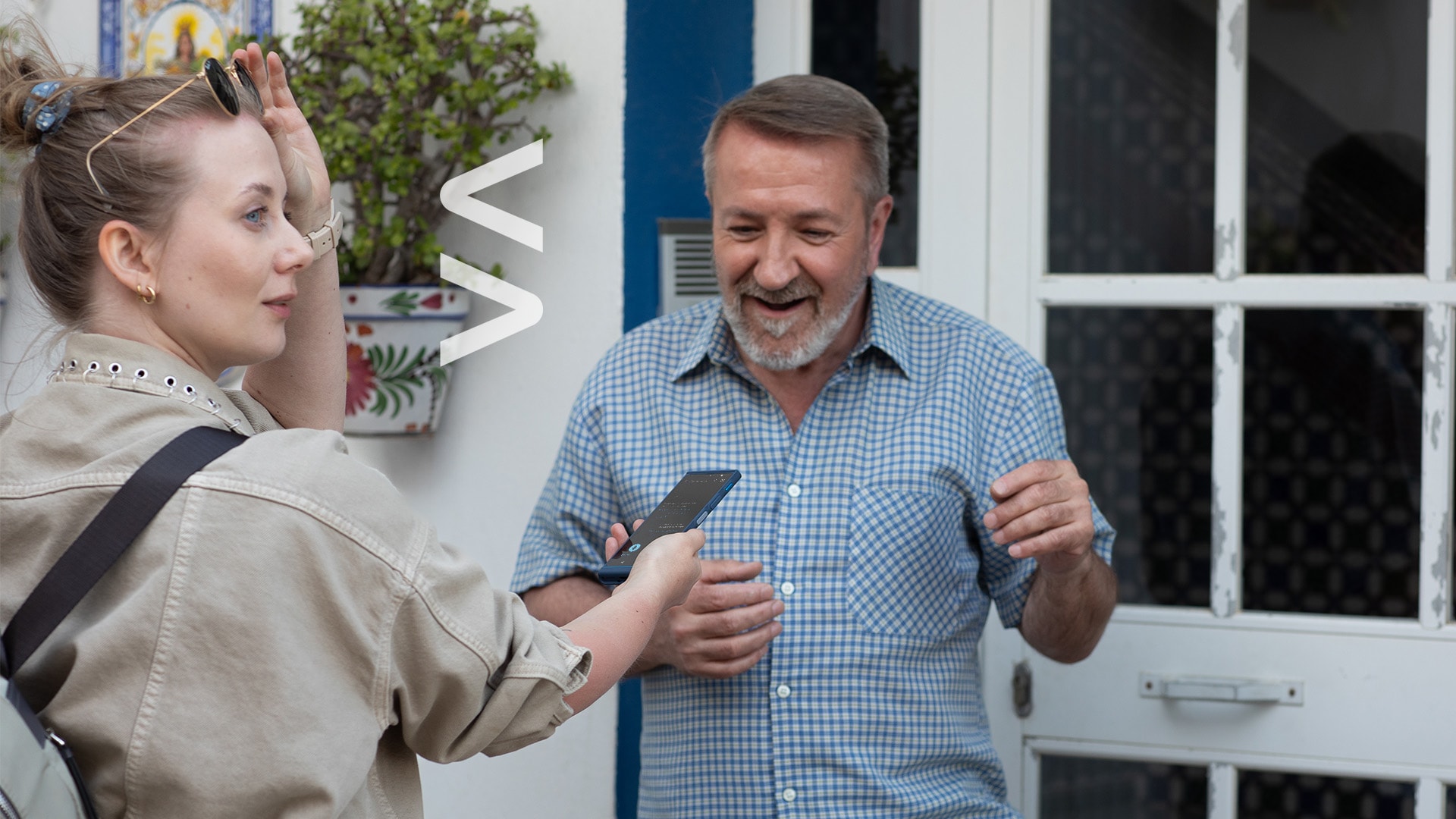 A woman holds a Vasco Translator V4 translator device while speaking to a smiling man outside a building, using it to assist with communication.