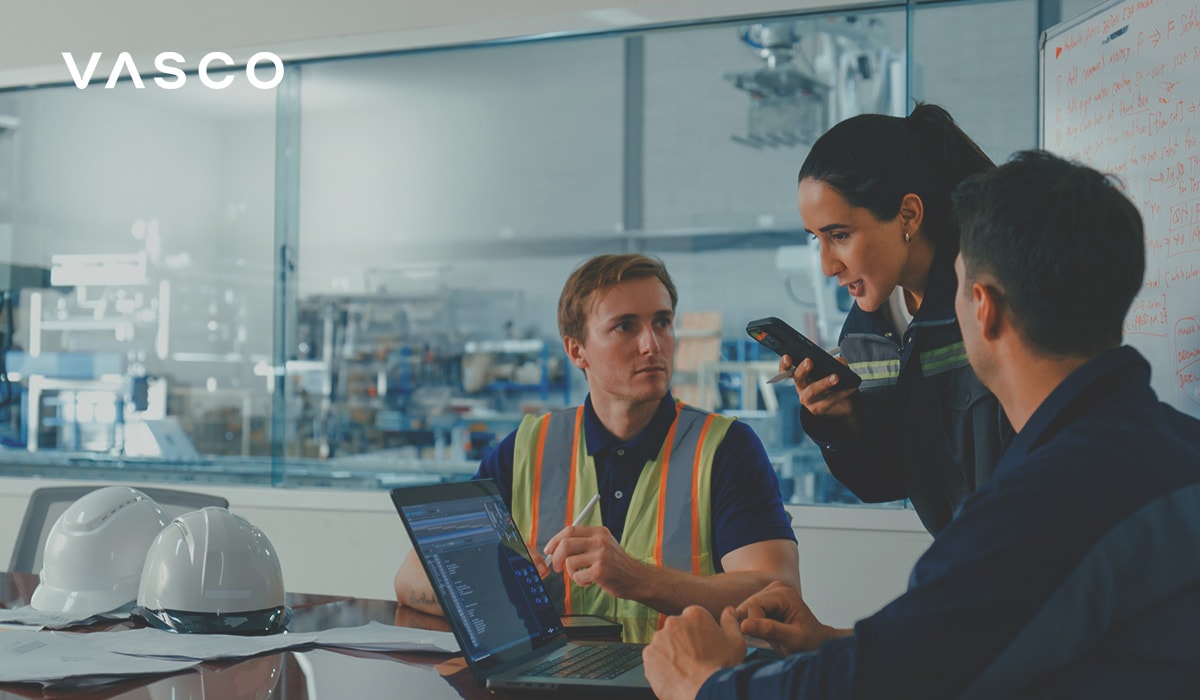 Workers in an industrial setting gathered around a table with laptops and documents. One person uses a smartphone for communication while others listen, with safety helmets on the table.