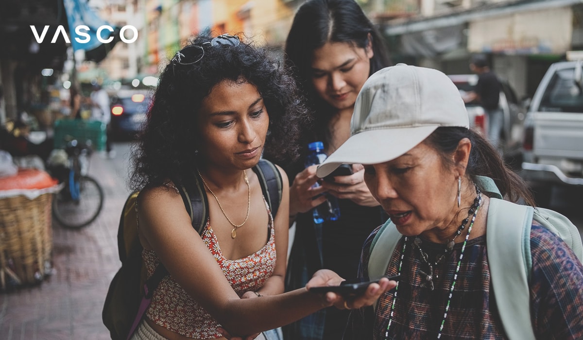 A group of three people standing on a busy street. One person holds out a smartphone, appearing to demonstrate or use a translation app, while the others observe attentively. The setting suggests a casual, real-world interaction in a multicultural environment.