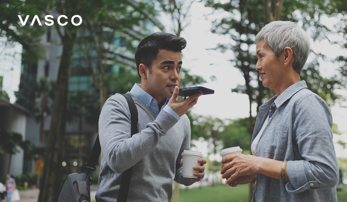 Two people conversing outdoors in a park or urban setting. A man holding a smartphone speaks into it, seemingly using a translation or voice feature, while a woman listens attentively, both holding takeaway coffee cups.