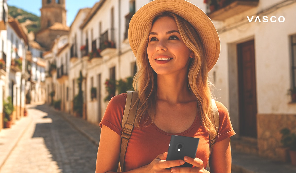 Smiling tourist woman walking through a narrow Spanish street with white buildings, holding a translation device and wearing a straw hat and backpack.