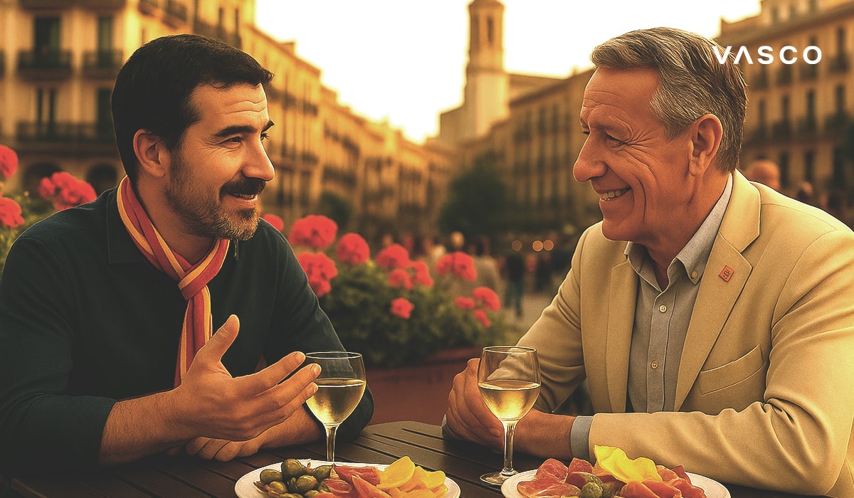 Two men enjoying drinks and tapas at an outdoor café in a Spanish square, engaged in conversation with flowers and architecture in the background.