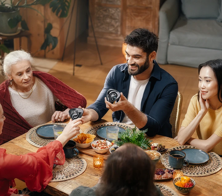 Multinational people gathered around a table, savoring a meal and engaging in lively discussions, creating a warm and inviting atmosphere while passing each other earbud Vasco Translator E1.