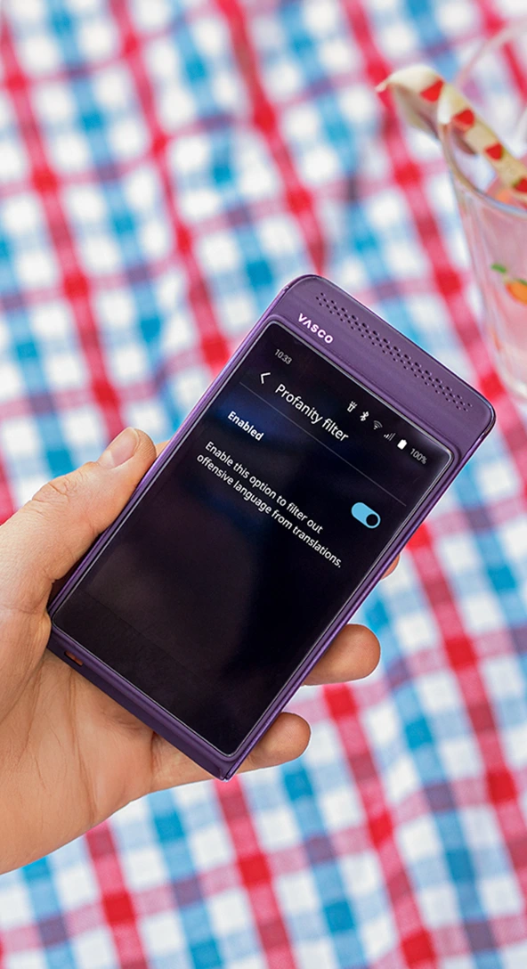 A person holds a purple Vasco electronic translator displaying the Profanity filter setting, with a checkered tablecloth and a glass of water with fruit slices in the background.
