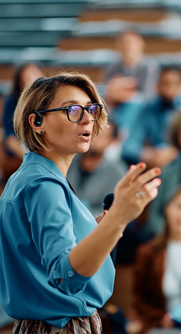 A woman wearing a Vasco E1 headset is speaking at a conference, with an audience of people visible in the background.