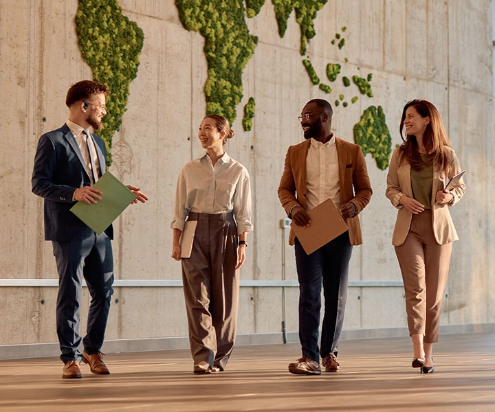 Four smiling people with briefcases walking through a modern office lobby, each wearing a Vasco Translator E1 in their ear. They appear relaxed and confident, engaged in conversation, with a professional and friendly atmosphere.