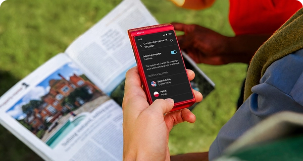 A person uses a red Vasco Translator Q1 to take a photo of a newspaper. The screen displays the language selection menu for translating the automatically detected text from the newspaper.